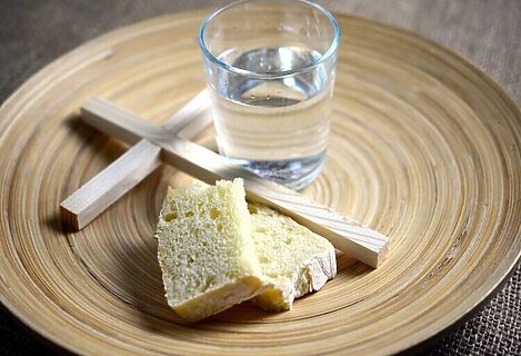 Wooden plate with cup of water, wooden cross, and crusty bread illustrate fasting